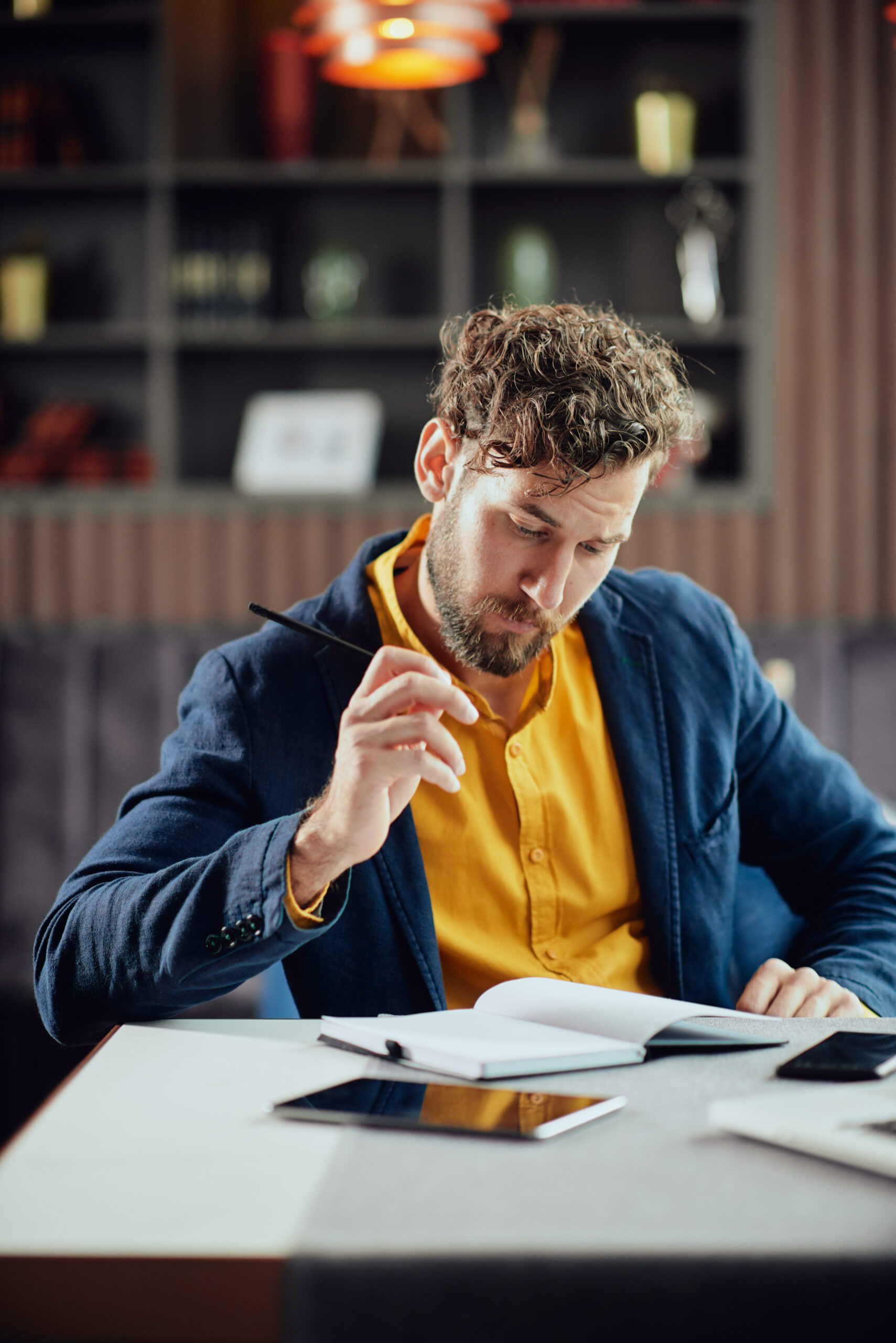 Young serious bearded Caucasian blogger dressed smart casual writing notes in agenda while sitting in cafeteria.