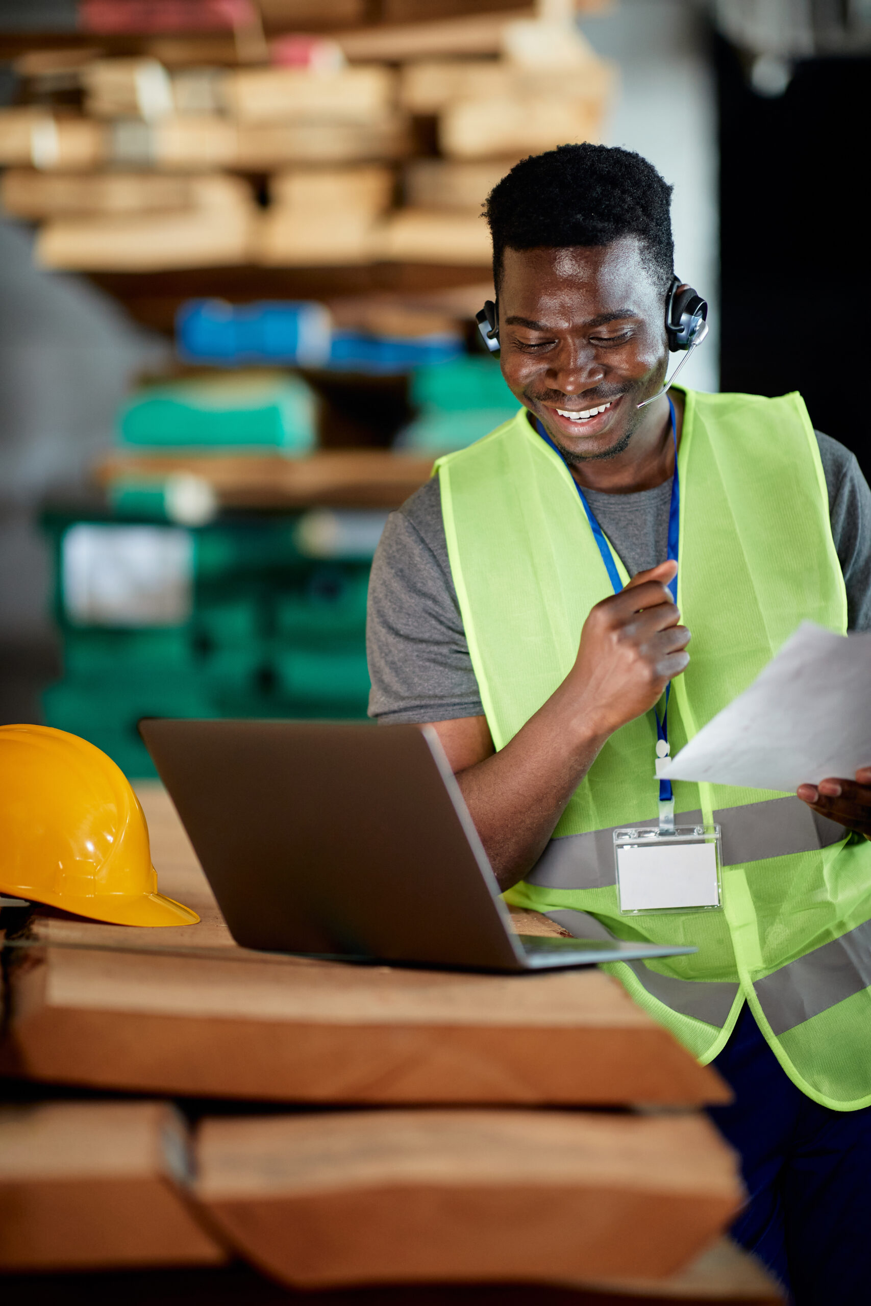 Happy black warehouse worker uses laptop and talks via headset while working at wood compartment,